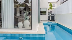 Et moderne udendørs swimmingpoolområde med et glasparti, et lille opholdsområde og en potteplante.