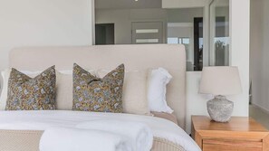 A bed with a beige headboard, two patterned pillows, a white pillow, and a wooden nightstand with a lamp. - Sandcastles (Casuarina)