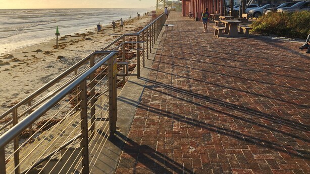 A paved walkway along a beach with a lifeguard tower, picnic tables, and a row of beachside buildings.