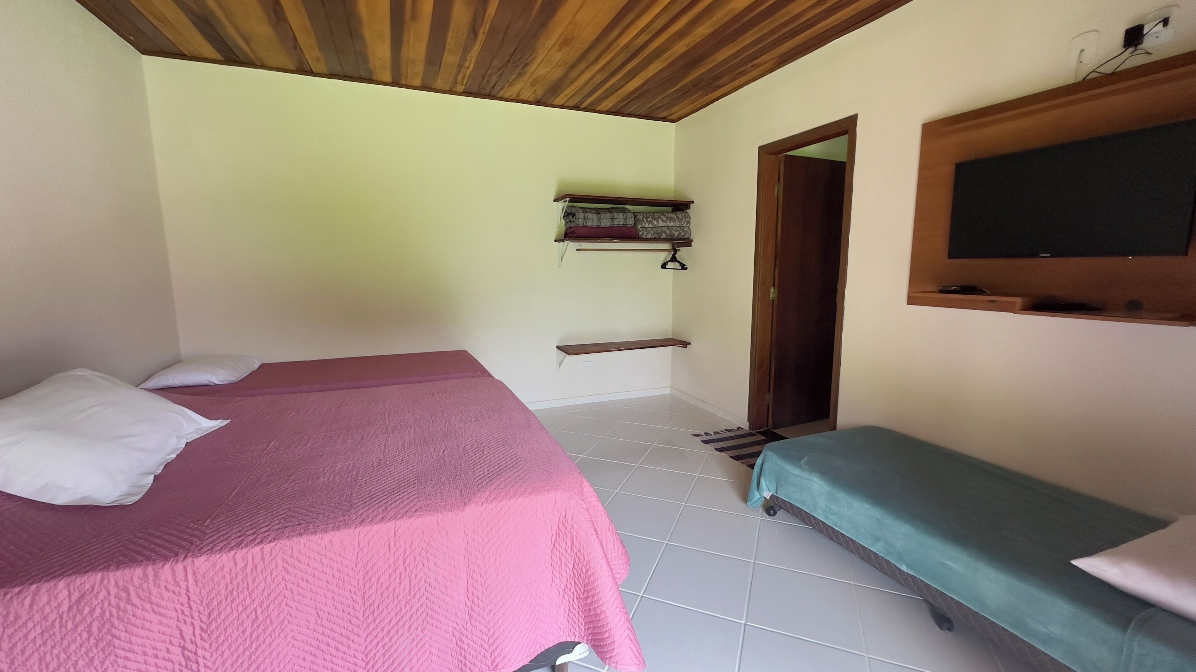 Una habitación con dos camas, una con cubrecama rosa y la otra con cubrecama verde. Tiene techo de madera y un estante con mantas dobladas.