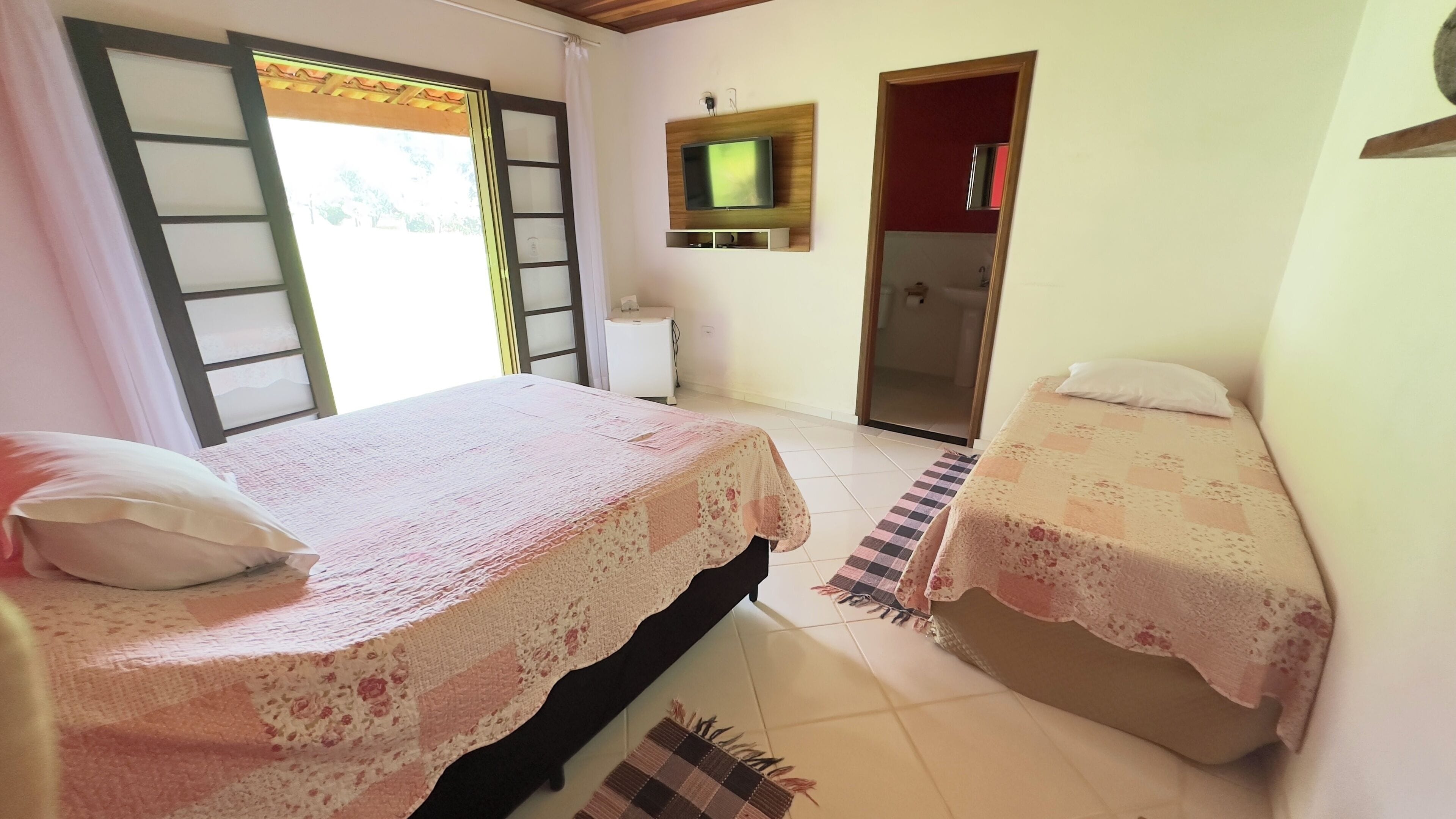 A bedroom with two beds, a TV mounted on the wall, and a bathroom visible through an open door.