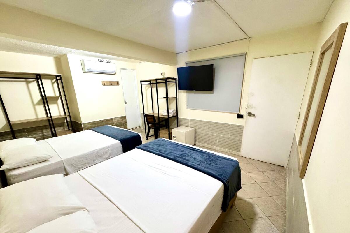 Ein Hotelzimmer mit zwei Einzelbetten, einem Schreibtisch mit Stuhl, einem an der Wand befestigten Fernseher und einer Tür.