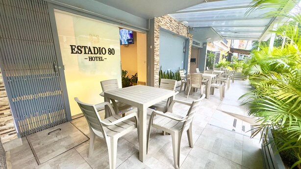 Un'area patio esterna con tavoli e sedie, un televisore montato a parete e un cartello con la scritta 'ESTADIO 80 - HOTEL'.