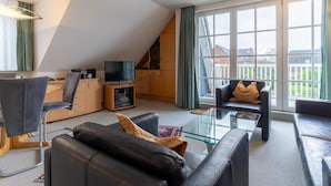 A modern living room with a glass coffee table, leather sofas, a television, and a view of a residential area through large windows. - Ferienwohnung in St. Peter-ording (St. Peter-Ording)