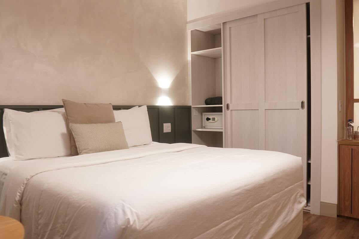 Ein ordentlich bezogenes Bett mit weißen Leinen und mehreren Kissen, ein Nachttisch, eine Mikrowelle und ein Holzschrank in einem modernen Raum mit Holzboden und hellen Wänden.