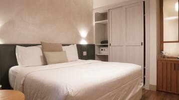 A neatly made bed with white linens and multiple pillows, a bedside table, a microwave, and a wooden cabinet in a modern room with a wooden floor and light-colored walls.