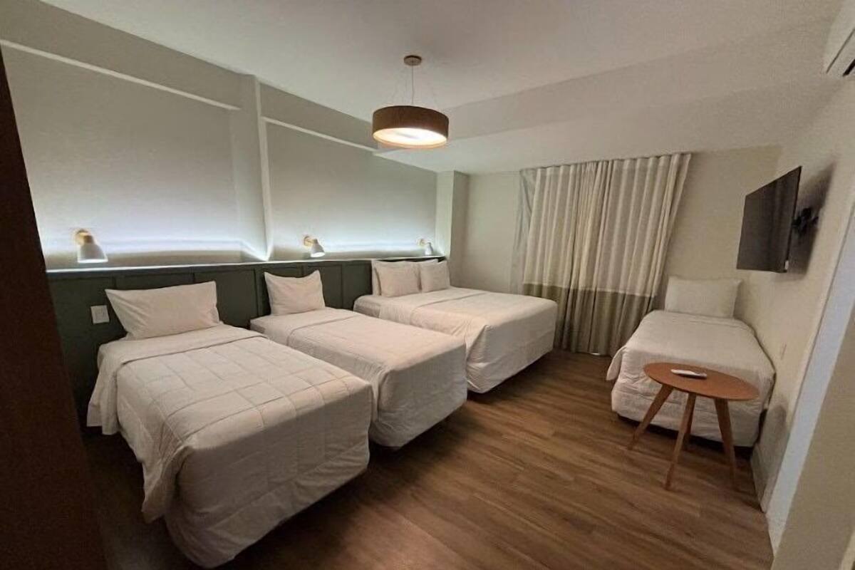 Ein Hotelzimmer mit drei Betten, einem kleinen Tisch, einem Fernseher und einem Fenster mit Vorhängen.