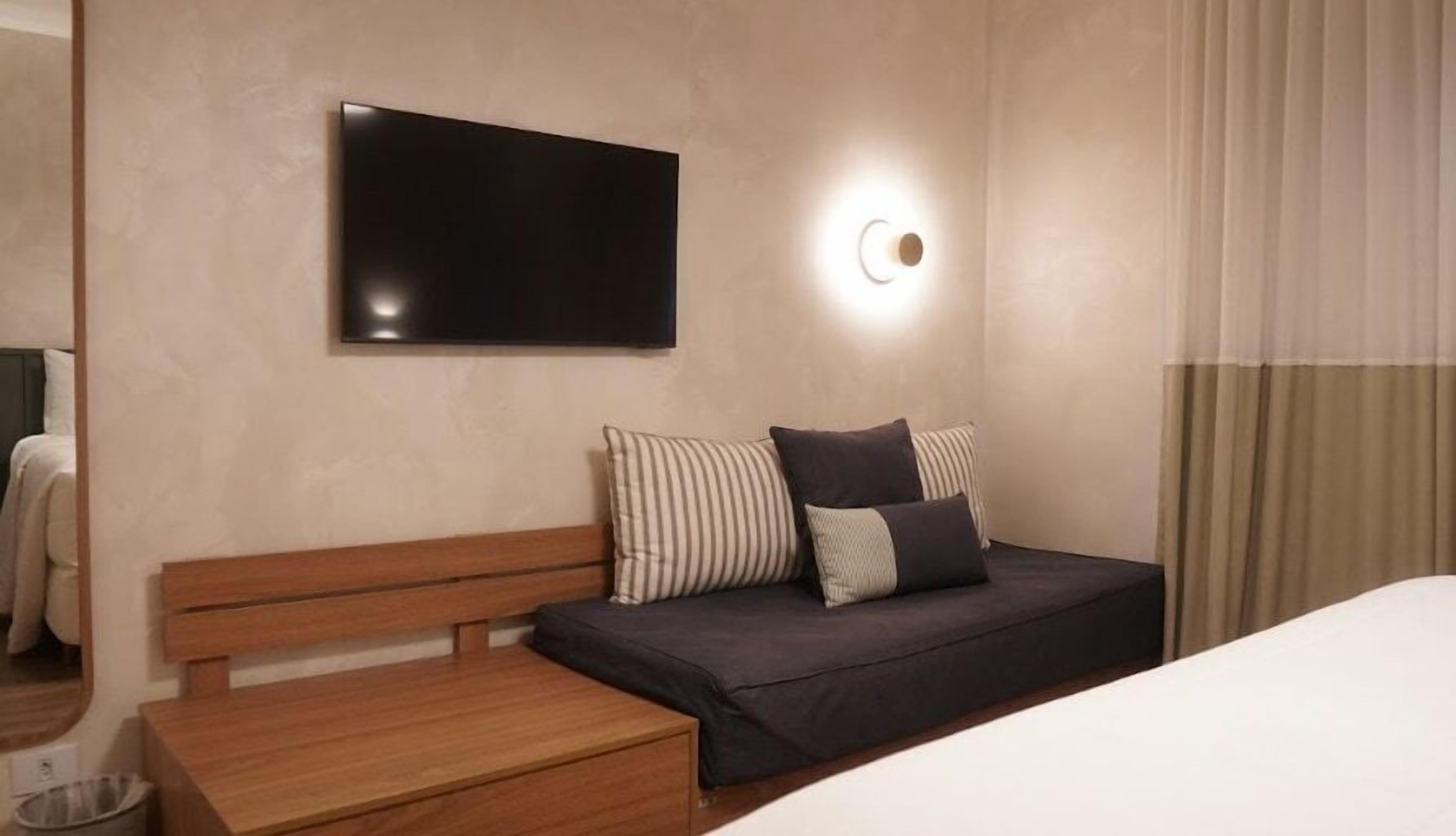 A modern hotel room with a wooden bench, a dark sofa with striped and solid pillows, a flat-screen TV, and a wall-mounted light.