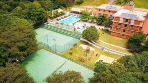 Vista aerea di un complesso sportivo con campo da basket, piscina e campo da tennis, immerso nel verde e con un edificio dotato di pannelli solari.