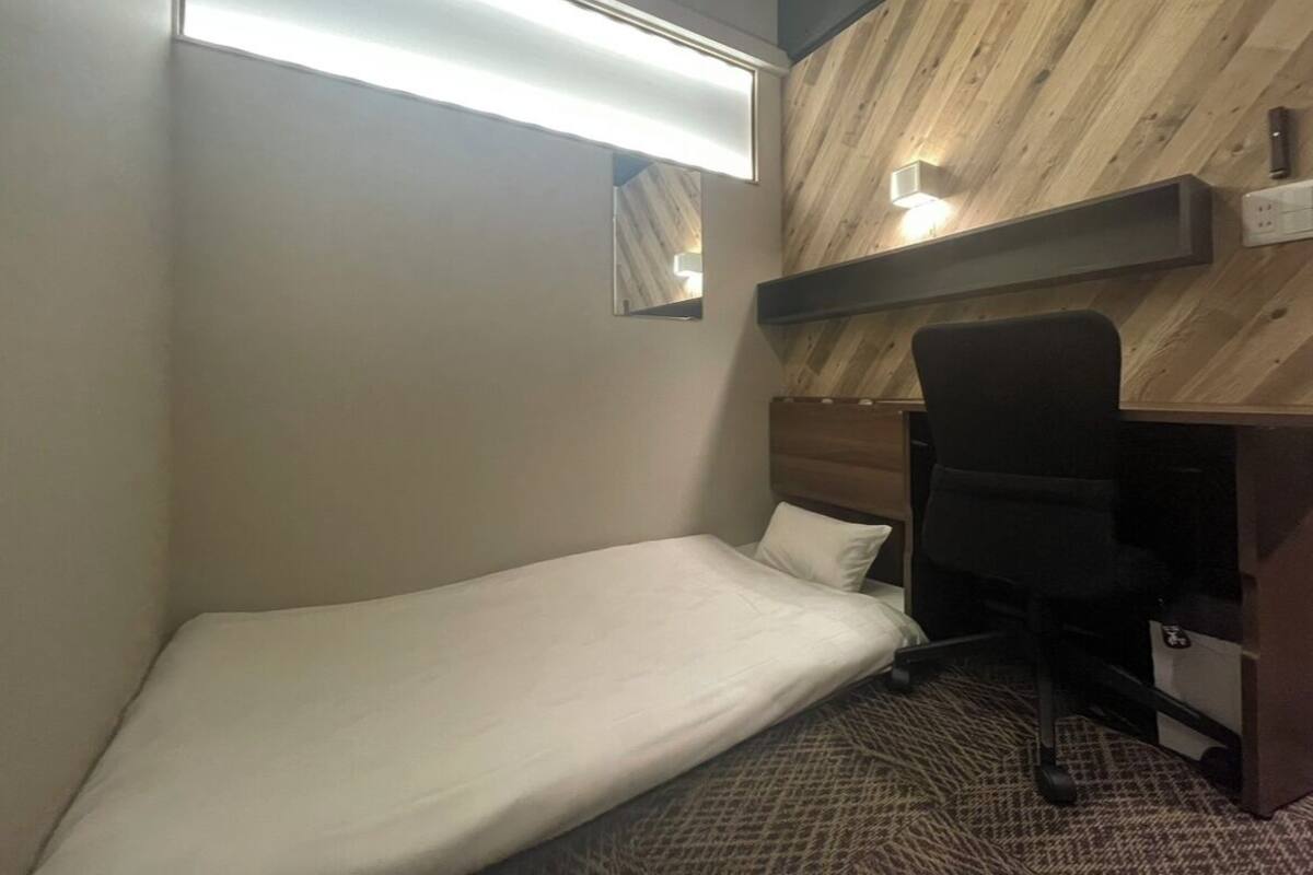 A small, compact room with a single bed, a desk, and a chair. There are two wall-mounted lights and a mirror above the bed.