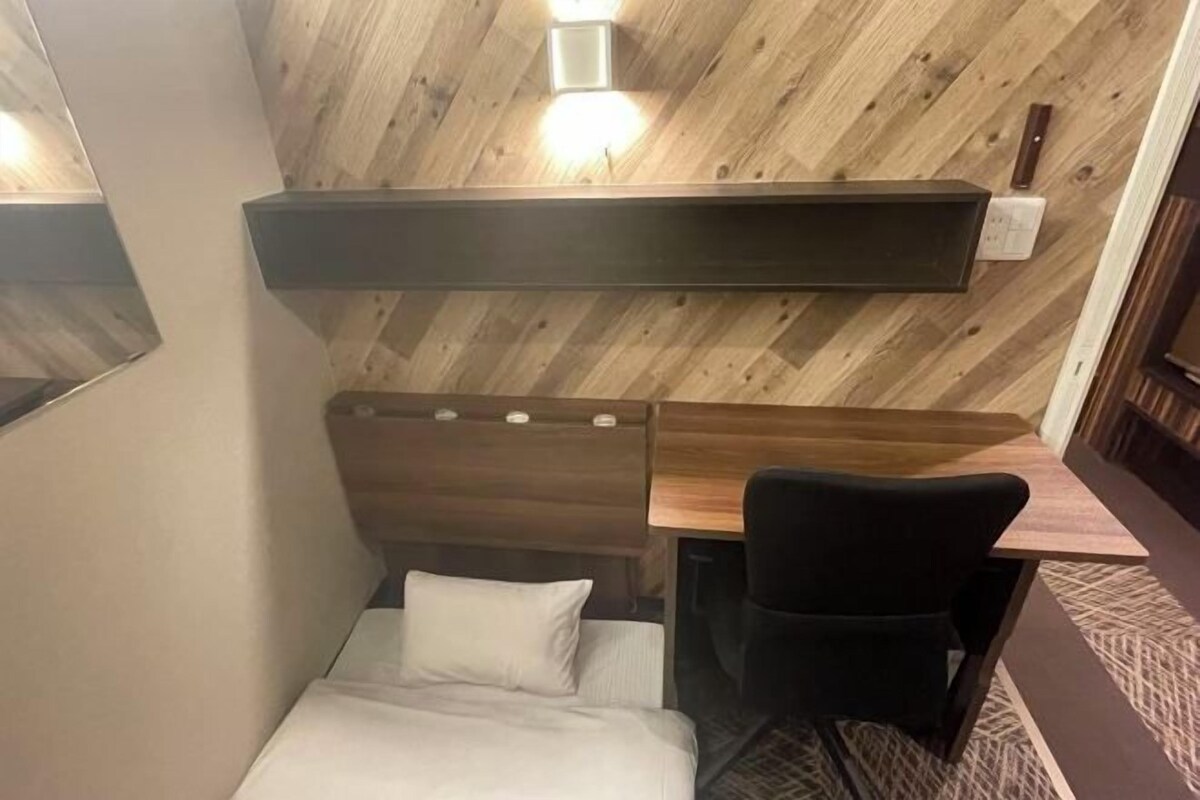 A compact hotel room with a wooden wall, a bed, a desk, a chair, and a small shelf.