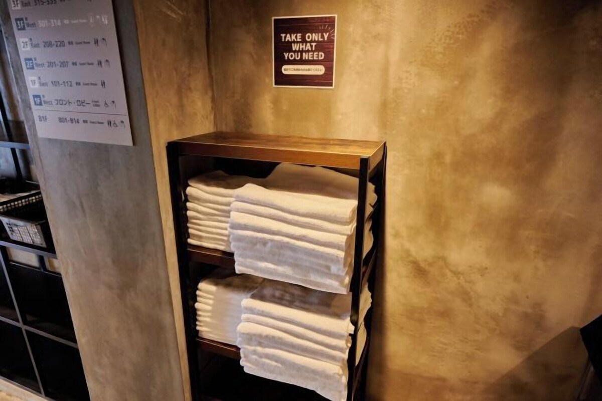 Una pila de toallas blancas sobre un estante de madera frente a una pared con textura, con un cartel que dice 'TAKE ONLY WHAT YOU NEED'.