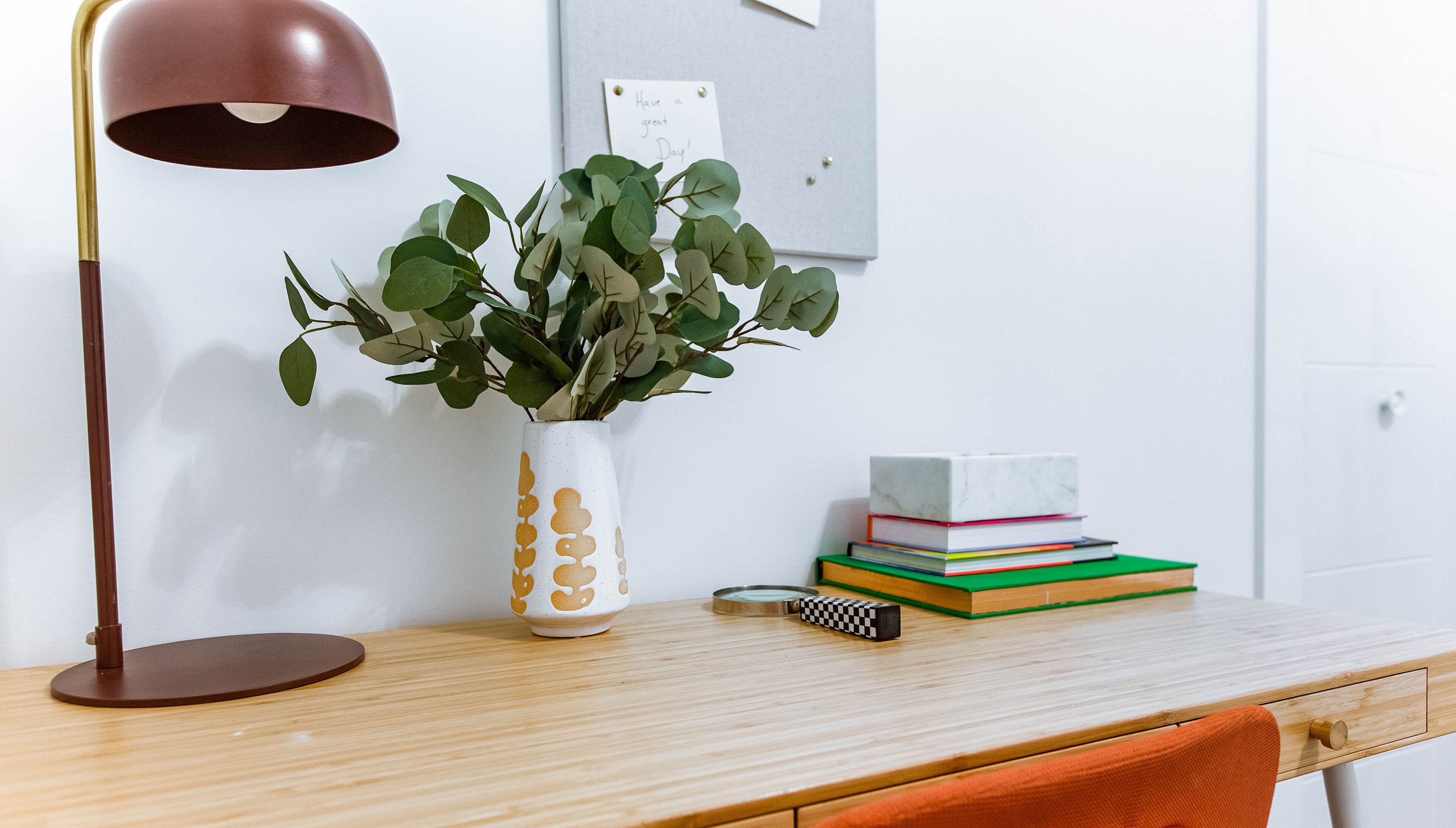 A wooden desk with a white vase containing green leaves, a stack of colorful books, a magnifying glass, and a brown desk lamp.