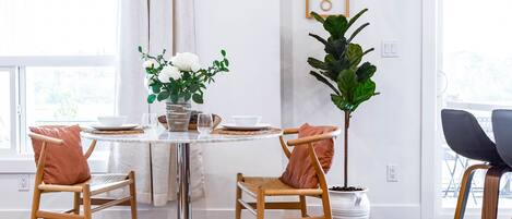 A dining area with a round table, four wooden chairs, a potted plant, a wall clock, and a view through a window.