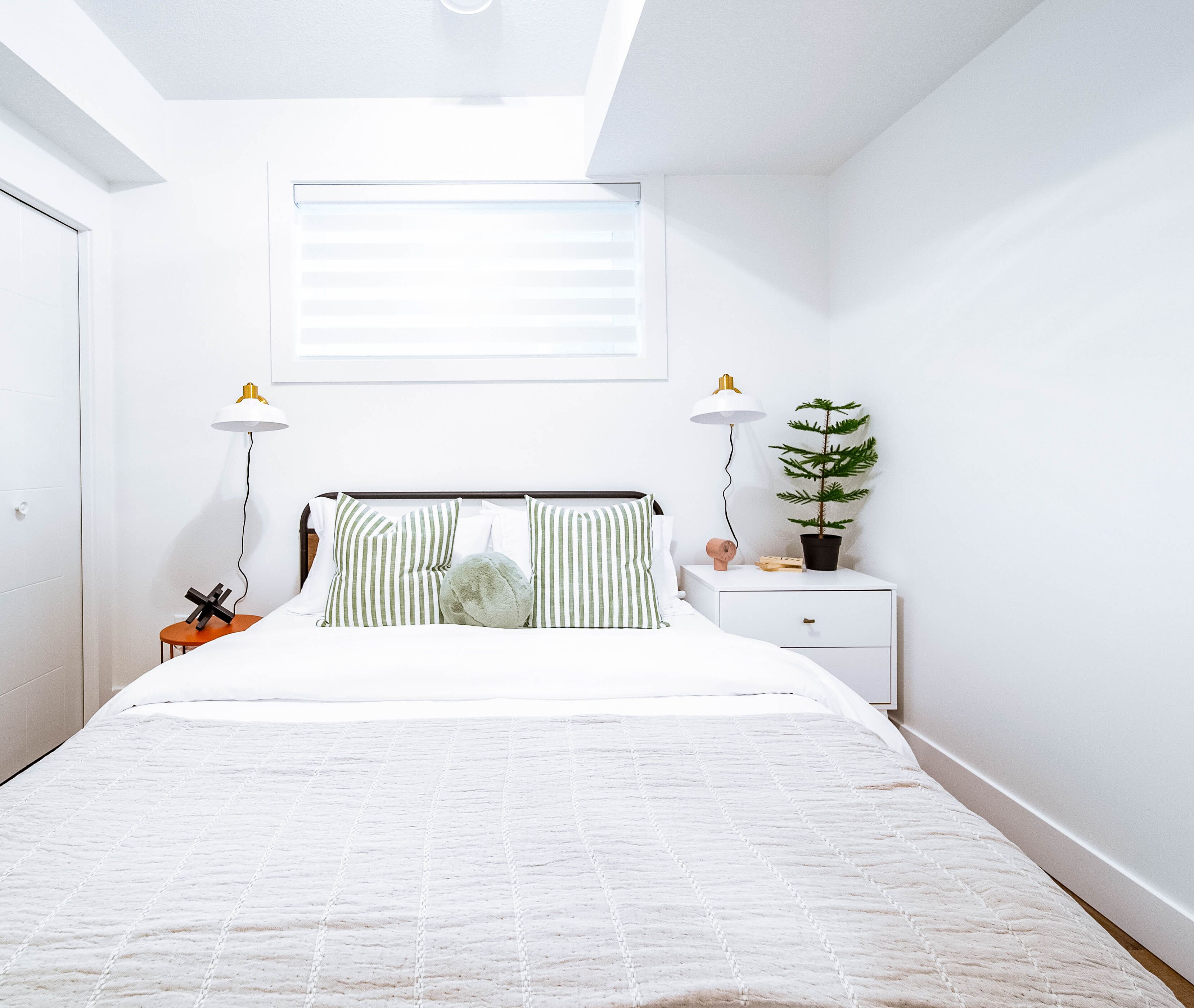 A bedroom with a bed, two bedside lamps, a nightstand with a plant, and a window with blinds.