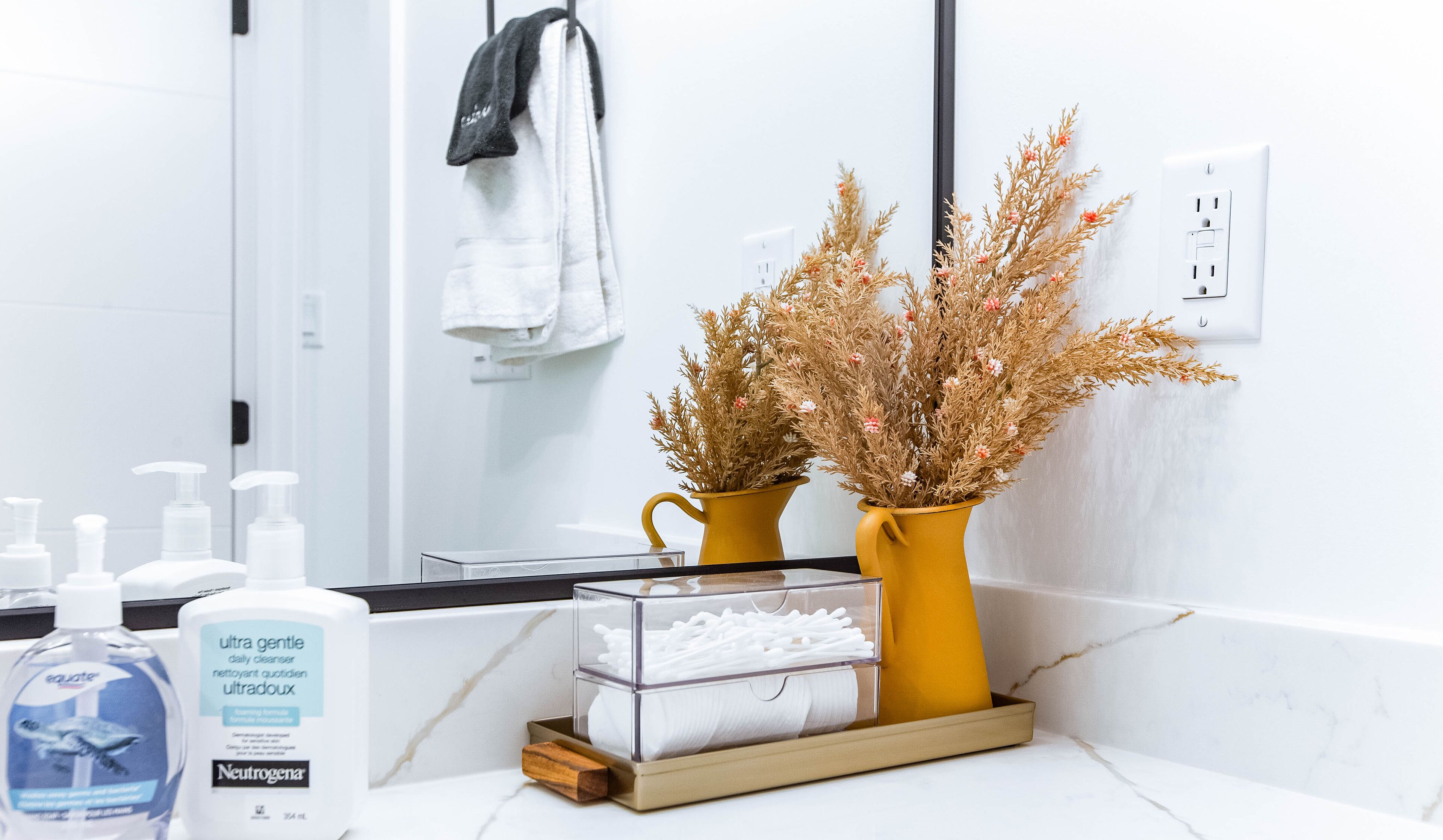 Een badkamer met witte tegels, een spiegel, een handdoekrek, een gele vaas met gedroogde bloemen, een zeepdispenser en een doosje wattenstaafjes.