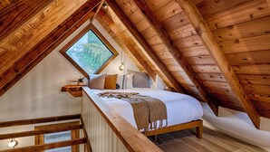 Un dormitorio acogedor en el ático, con techo inclinado de madera, un ventanal grande, una cama con cubrecama blanco y un banco de madera.