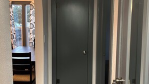 Un passadís amb una porta gris fosca, una zona de dinar amb taula i cadires, i una finestra amb cortines estampades.
