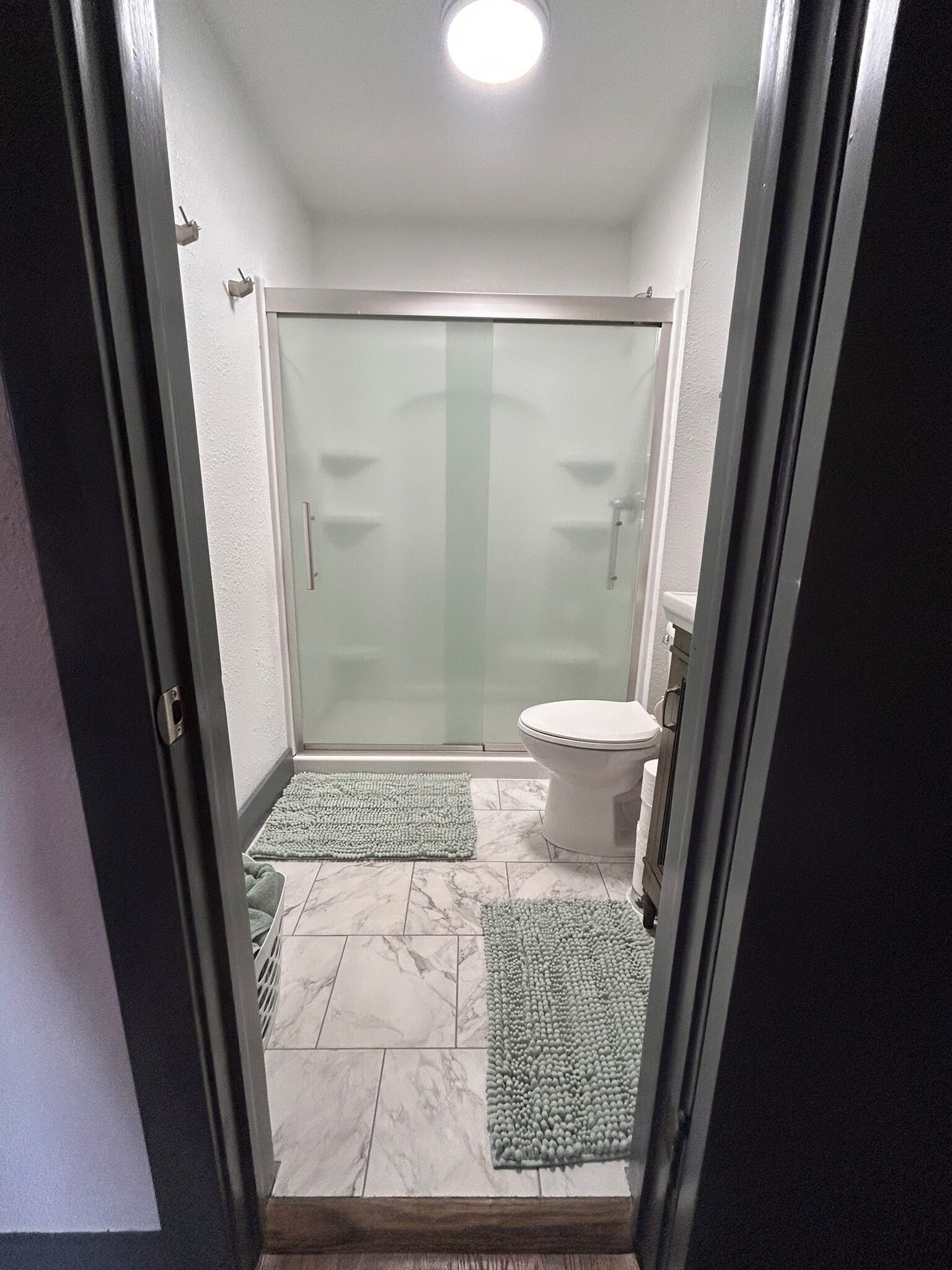 A bathroom with a glass shower door, a toilet, and two green mats on the floor.