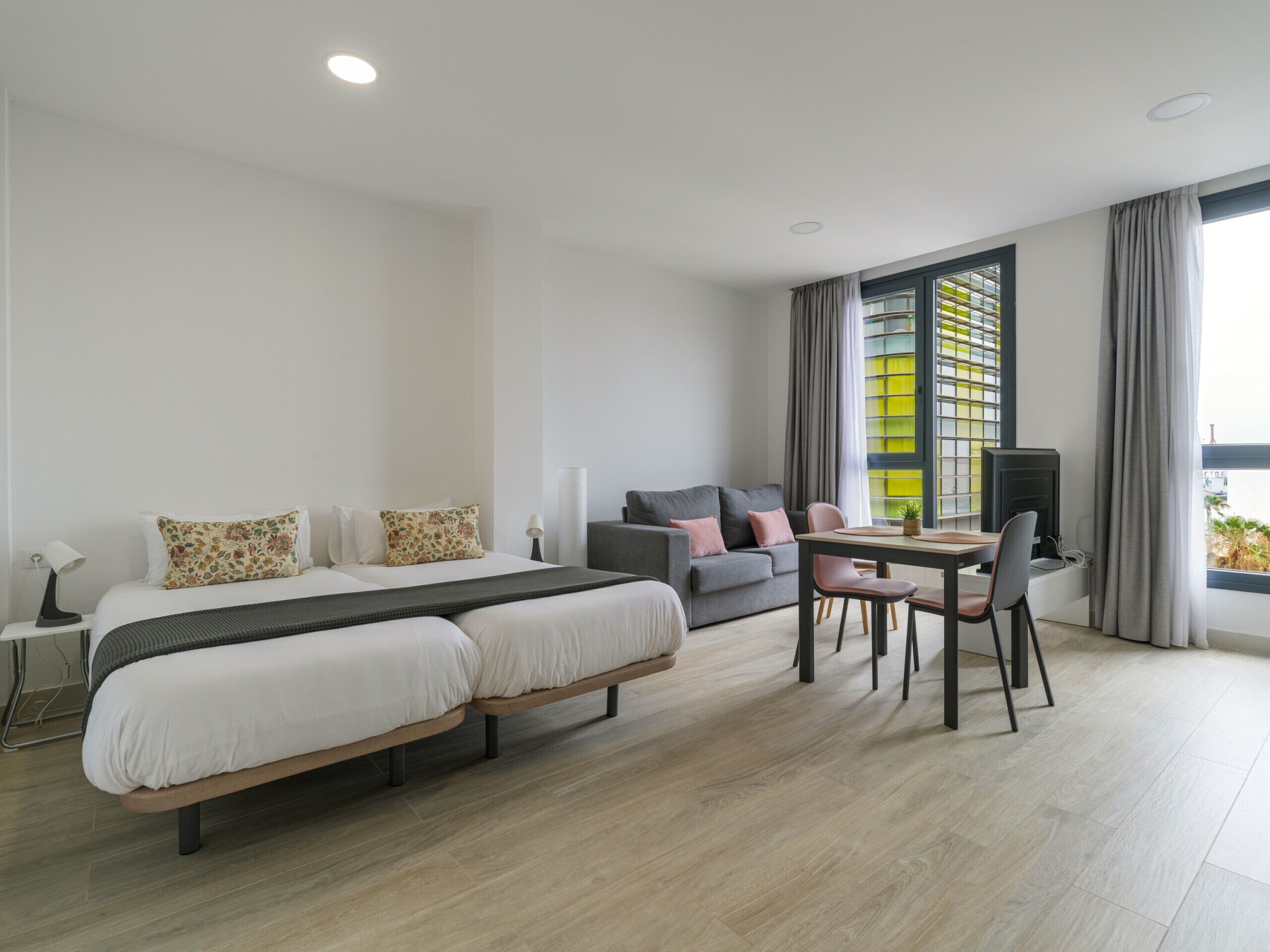 Une chambre d’hôtel moderne équipée d’un lit double, d’un canapé, d’une table à manger avec des chaises, d’une télévision et d’une grande fenêtre avec des rideaux.