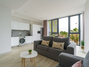 A modern apartment with a grey sofa, a small round table, a washing machine, and a kitchen area.