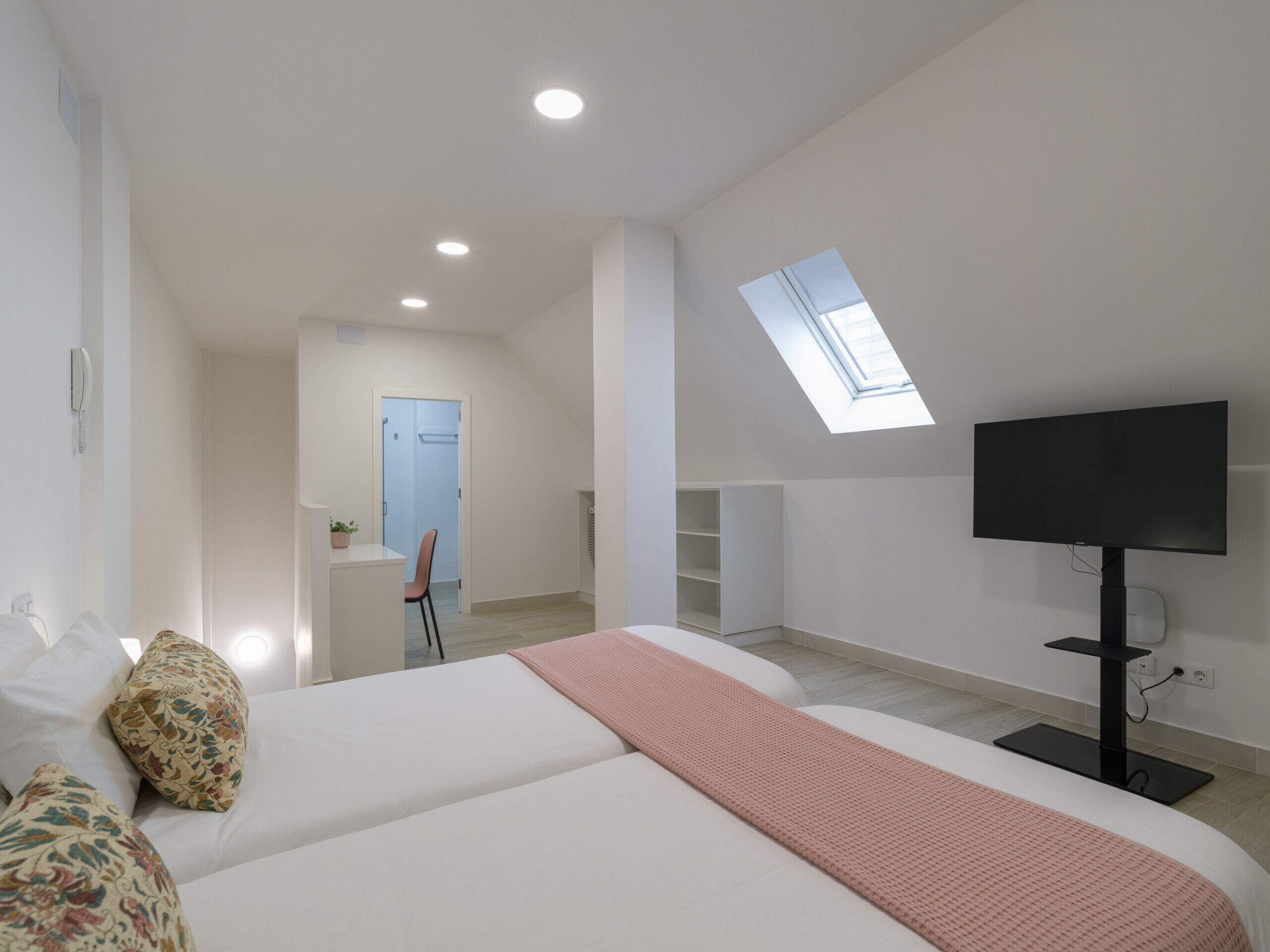 Un dormitorio moderno con una cama grande, televisión, tragaluz y una pequeña zona de escritorio.