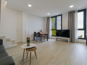 A modern living room with a flat-screen TV, a dining area with chairs, a staircase, and a window with curtains.