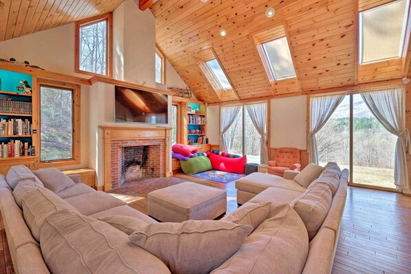 A spacious living room with a fireplace, large windows, and a high ceiling.