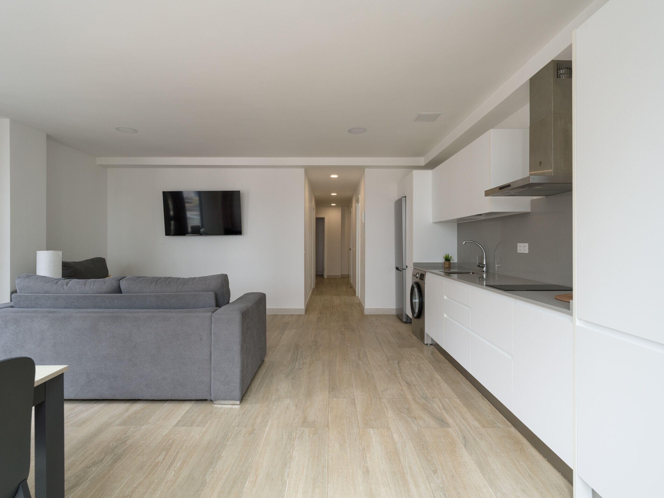 A modern living room with a grey sofa, a flat-screen TV, and a washing machine in the background.