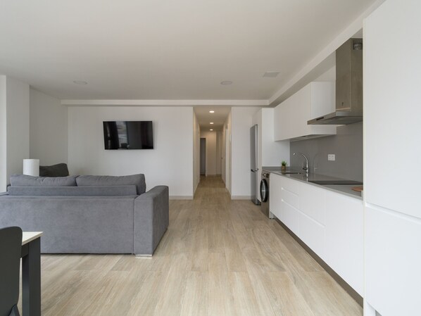 A modern living room with a grey sofa, a flat-screen TV, and a washing machine in the background.