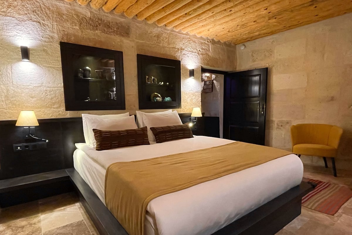 A bedroom with a large bed, two bedside tables with lamps, a wooden ceiling, stone walls, and a door.