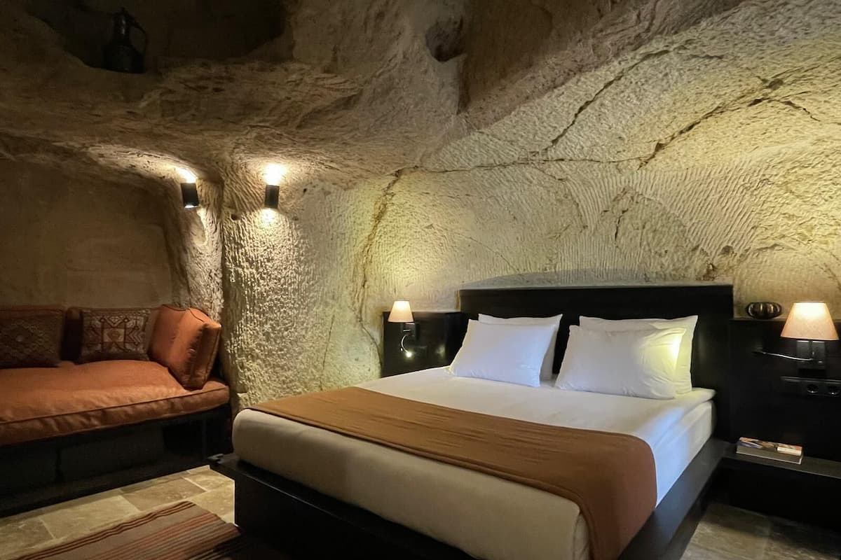 A bedroom with a stone ceiling, a bed with white linens, a brown sofa, and two bedside lamps.