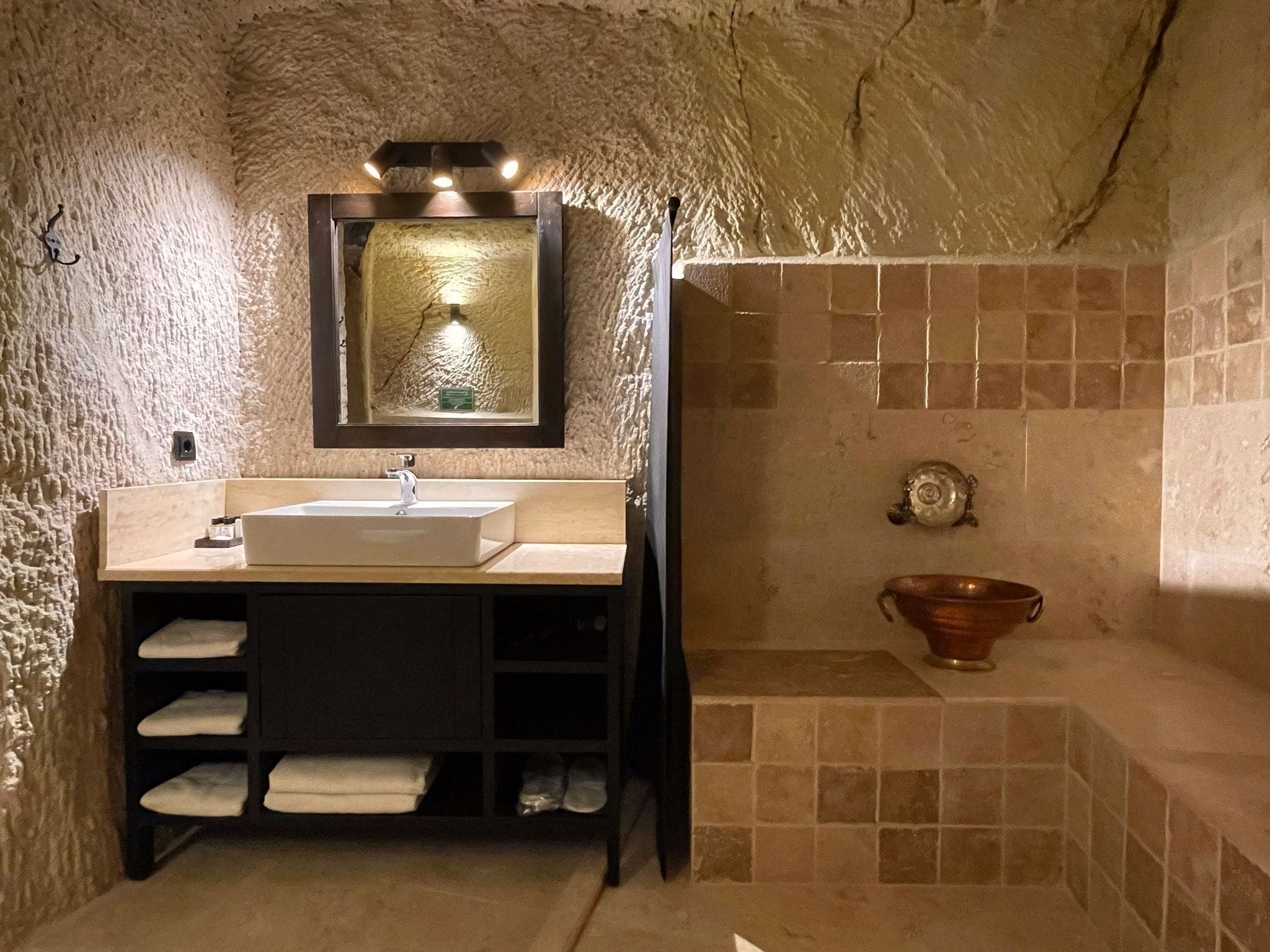 A bathroom with a stone wall, a sink with a mirror, a towel rack, and a bathtub with a decorative bowl.