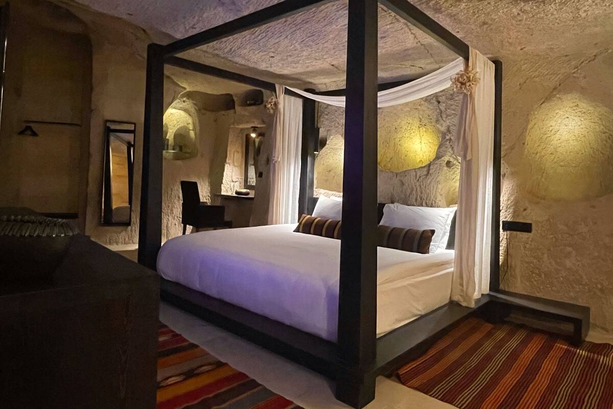 A four-poster bed with white linens, a striped rug, and a stone wall.