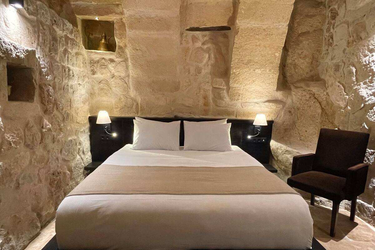A bed in a room with stone walls, two bedside lamps, and a chair.