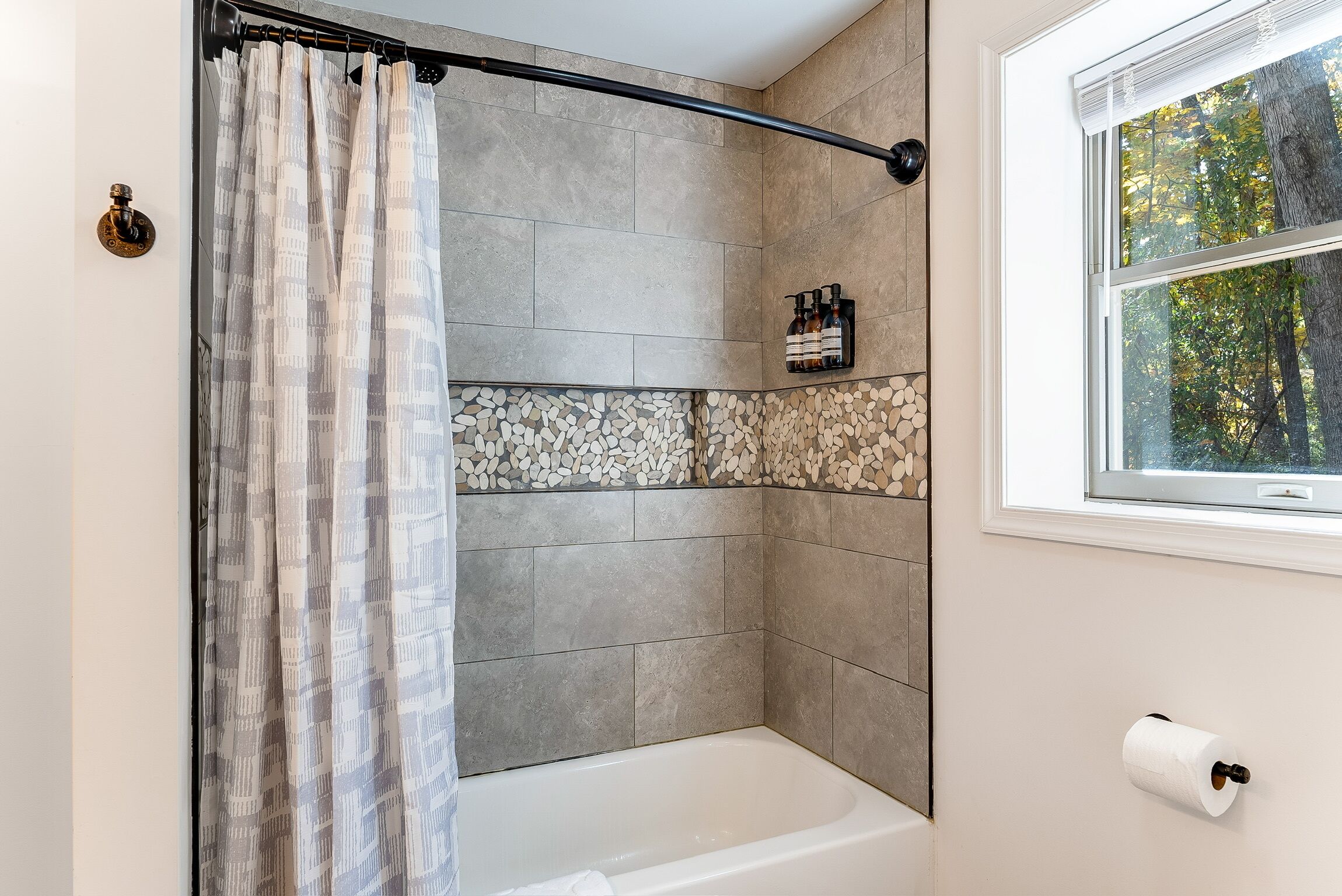 Ein Duschbereich mit Vorhang, eine Badewanne, ein Fenster mit Blick auf Bäume und ein Toilettenpapierhalter.