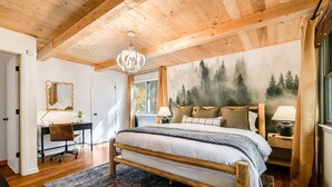 Een slaapkamer met een houten bed, een bureau, een stoel, een groot raam en een bosdecoratie op de muur.