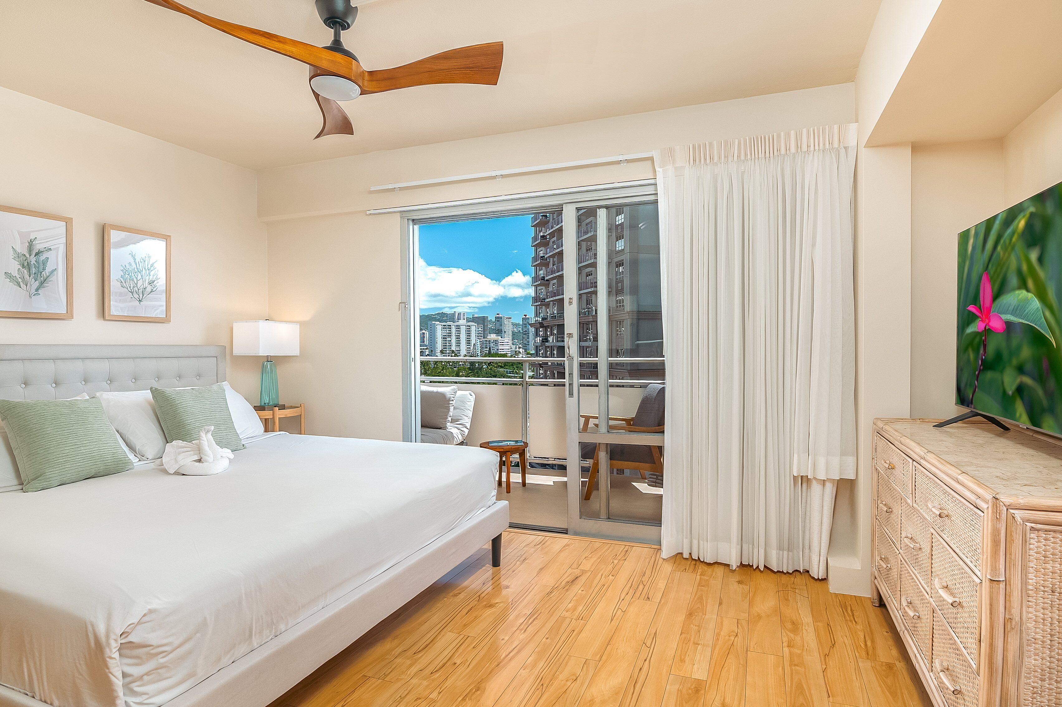 Une chambre avec un grand lit, un ventilateur de plafond, une télévision, un balcon avec vue sur les immeubles et un plancher en bois.