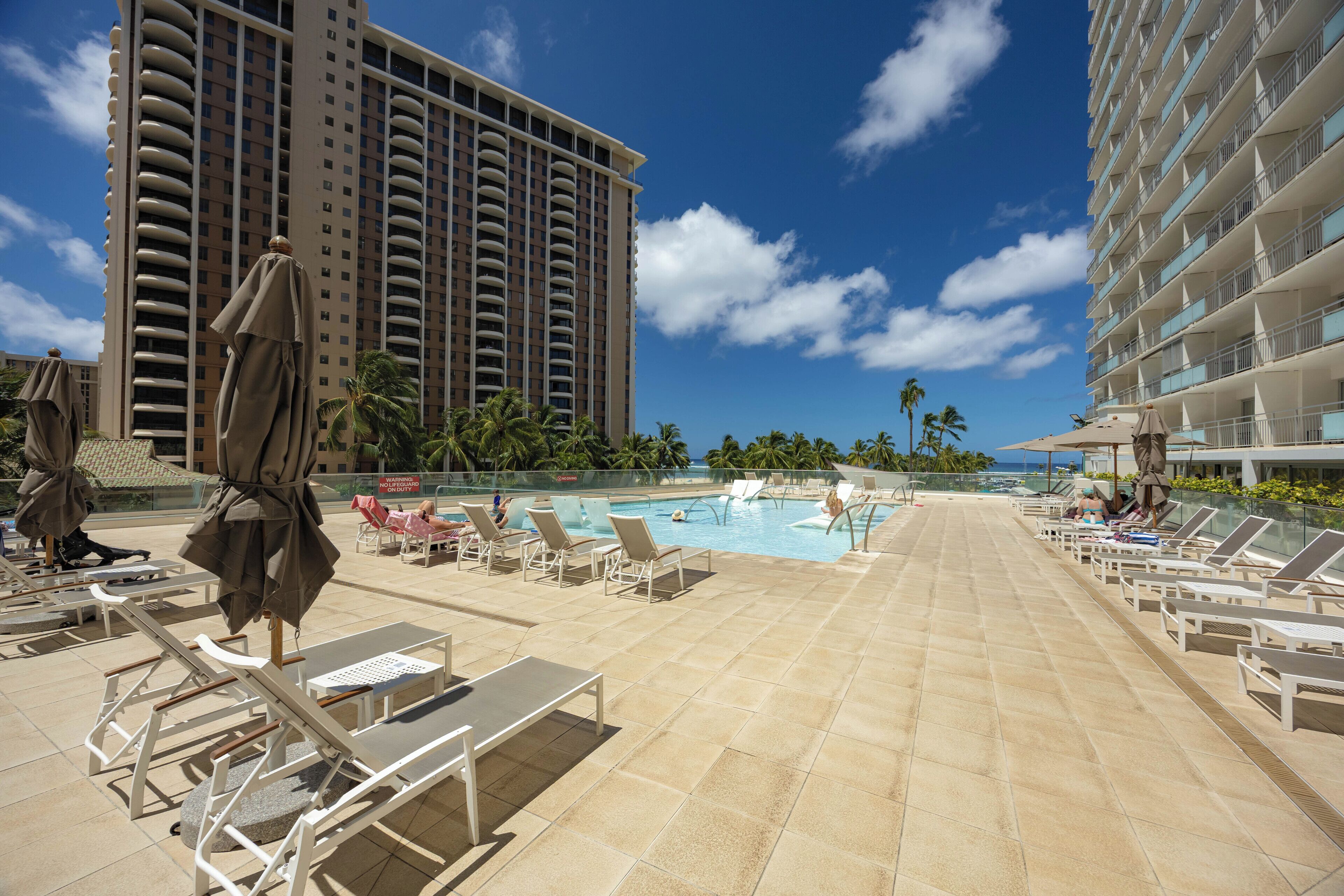 A hotel pool area with sun loungers and umbrellas, a clear sky, and a tall building in the background.