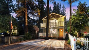Une maison en bois à deux étages, dotée d’un balcon, entourée de grands arbres et d’une clôture blanche en piquets.