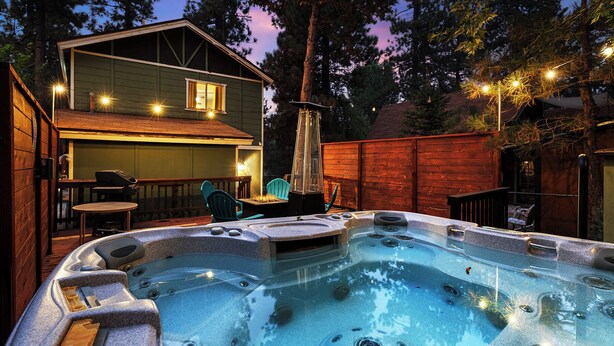 Un jacuzzi dans un jardin, avec une terrasse en bois, un espace barbecue et une maison dotée d’un toit en pente.