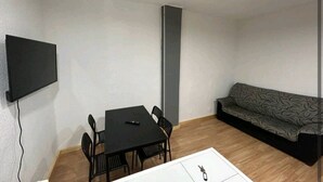 Une petite pièce moderne, avec une table à manger noire, quatre chaises, un canapé gris et un téléviseur à écran plat fixé au mur.
