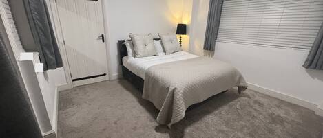 Un dormitorio con cama, una puerta, ventana con persianas y suelo cubierto de alfombra.