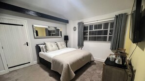 Un dormitorio con cama, una mesita de noche con lámpara, ventana con cortinas, un espejo y un televisor en la pared.