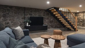A modern living room with a stone wall, a black entertainment unit, a blue sofa, and a wooden staircase. - Loki’s Loft and Cellar (Kaslo)