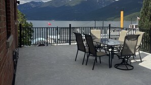 A patio with a table and chairs, a view of a lake, mountains, and a flag. - Loki’s Loft and Cellar (Kaslo)