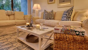 Una sala de estar acogedora con dos sofás color beige, una mesa de centro blanca con velas y vasos, una cesta tejida y una caja de juego de mesa.