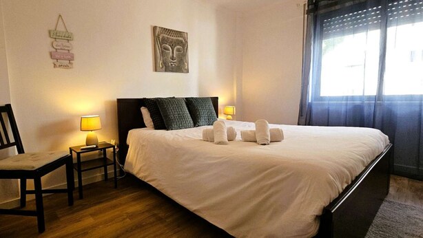 A hotel room with a bed, a chair, a bedside table, a wall decoration, and a window with curtains.