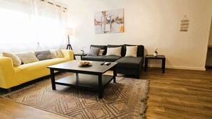 A living room with a yellow sofa, a black sofa, a coffee table, a patterned rug, and a painting on the wall.