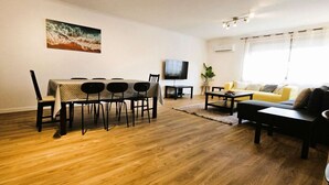 A living room with a wooden floor, a dining table with chairs, a sofa, a TV, and a plant.